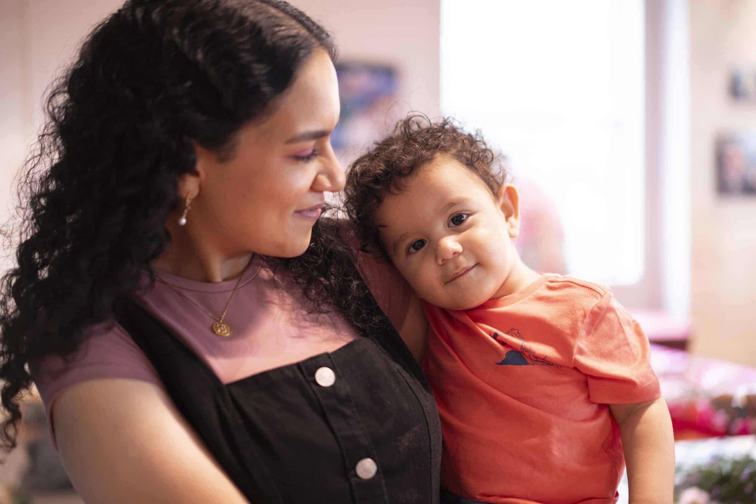 Image of mother holding young child. Mother is looking at her child. Child is looking at the camera.
